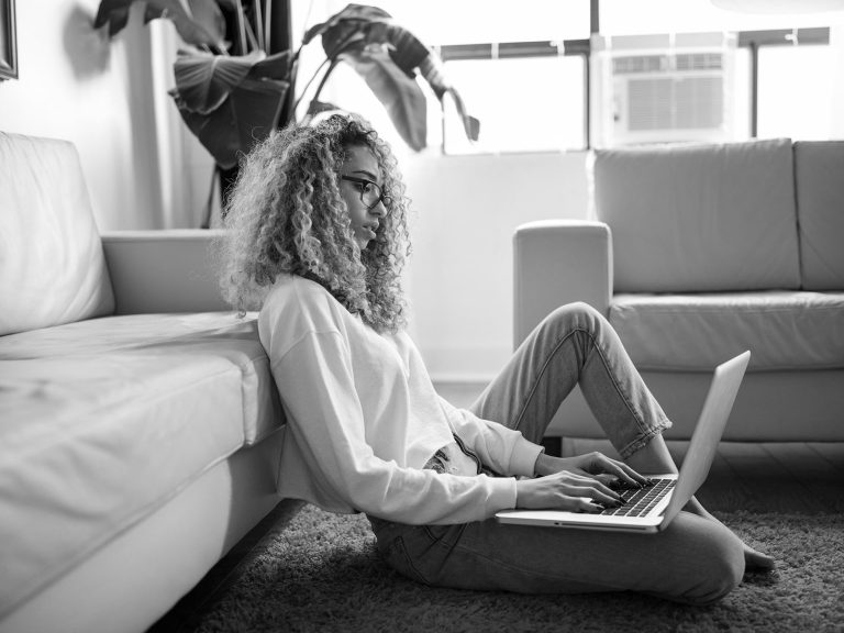 Frau mit lockigem Haar sitzt auf dem Boden und arbeitet am Laptop.